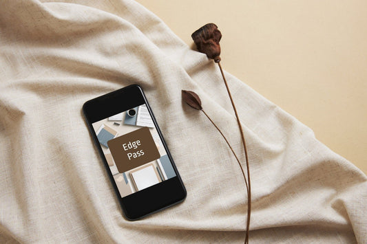 A black smartphone lying on draped beige fabric, displaying a digital pass that reads 'Edge Pass'. A dried brown flower stem rests beside it on a warm beige background.