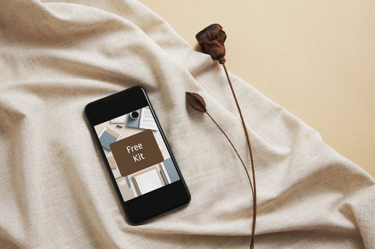 A black smartphone lying on draped beige fabric, displaying a mockup screen with a brown card labeled 'Free Kit.' A dried brown flower rests beside it on a warm beige background.