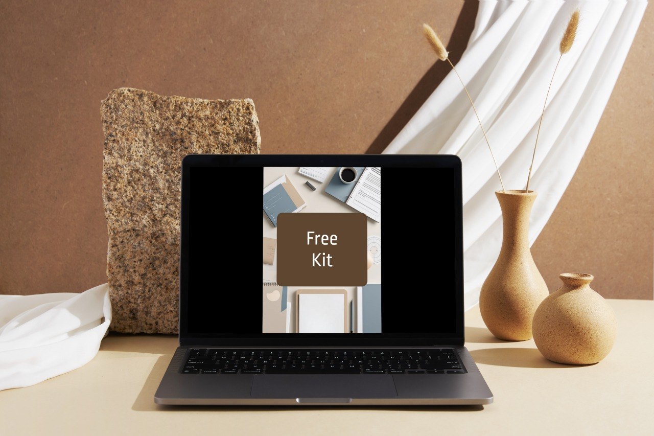 A dark gray laptop sits open on a beige tabletop, displaying a mockup with the text 'Free Kit' on the screen. The scene is styled with a textured stone block, white draped fabric, and two beige ceramic vases with dried stems against a warm brown wall.