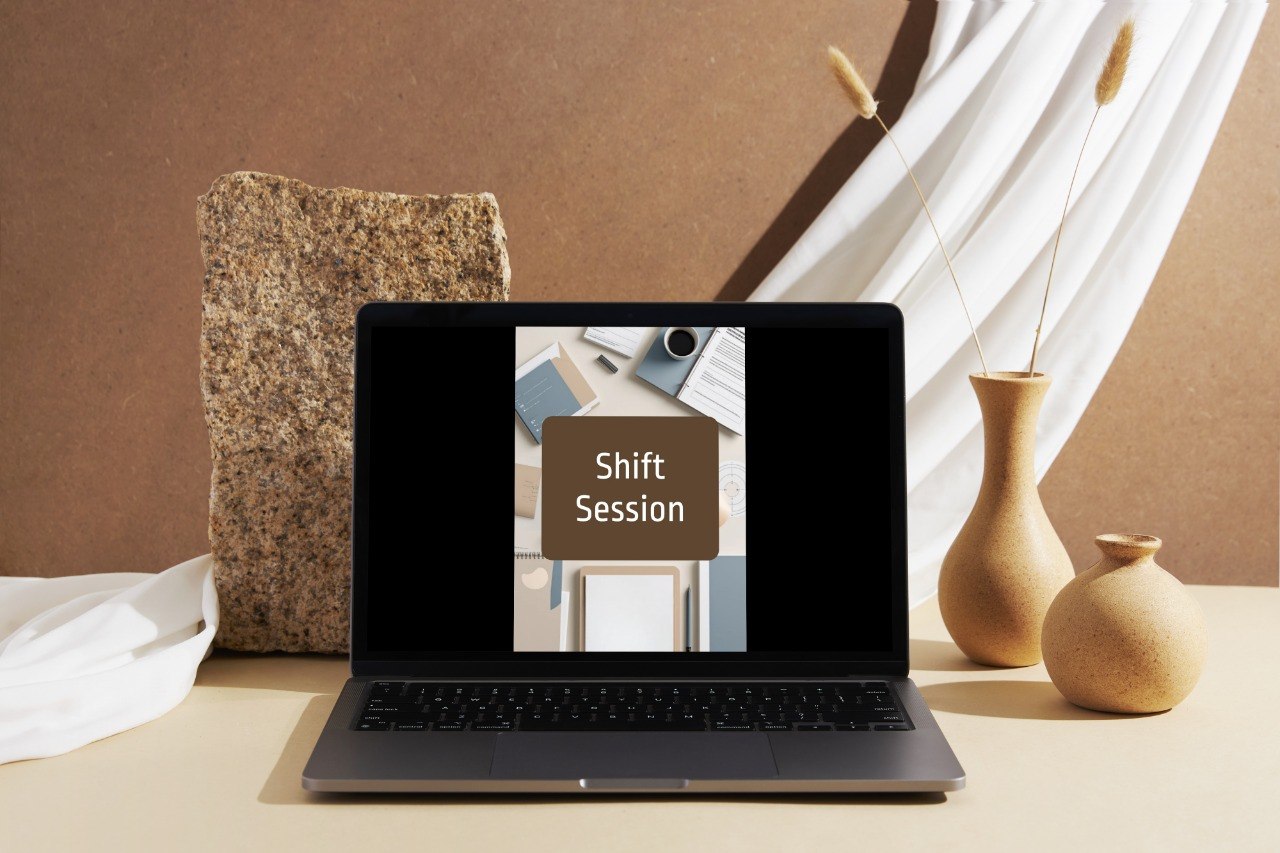 A dark gray laptop sits open on a beige tabletop, displaying a digital course or meeting screen that reads 'Shift Session.' The scene is styled with a brown textured wall, draped white fabric, a stone slab, and two beige ceramic vases in the background.
