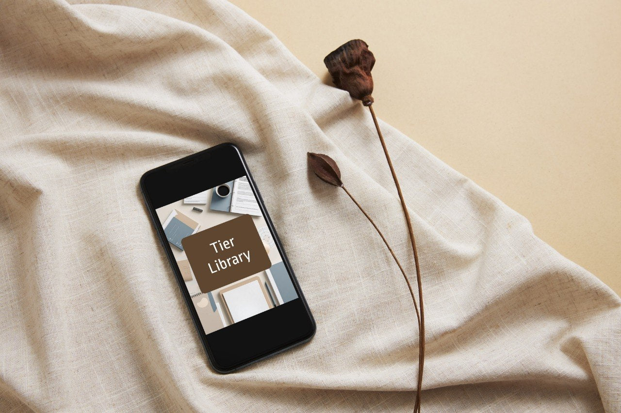 A black smartphone lying on draped beige fabric, displaying a book cover that reads 'Tier Library.' A dried brown flower stem rests beside it on a warm beige background.